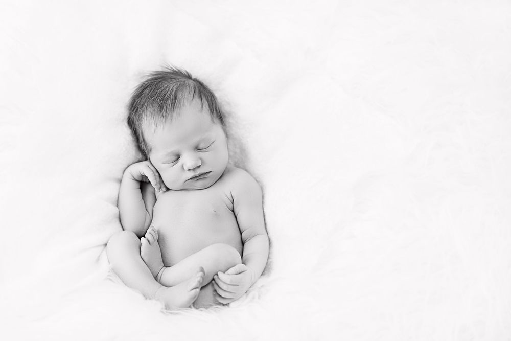 A sleeping newborn baby curled up peacefully on soft white fabric in black and white.