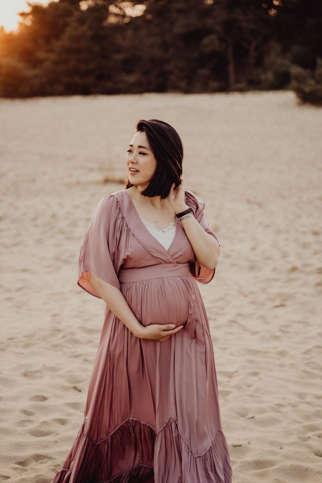 A maternity portrait on sandy beach wearing flowing mauve dress at sunset.
