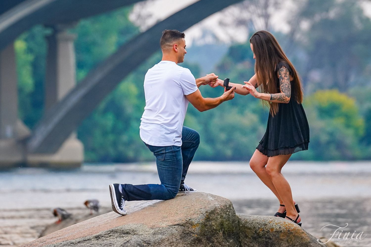 A joyful marriage proposal takes place on a rocky riverbank with a bridge in the background.