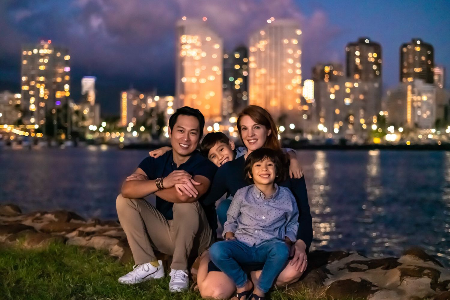 A family sitting together on waterfront with illuminated city skyline in background at dusk. Family Portrait Photography