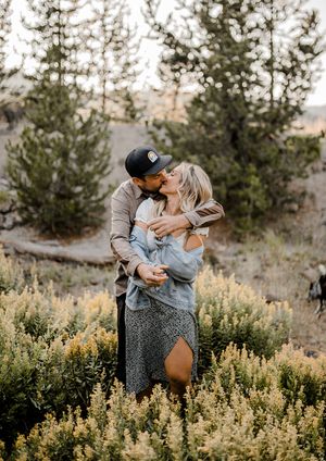 Romantic couple embracing in a field of wildflowers surrounded by pine trees at golden hour.