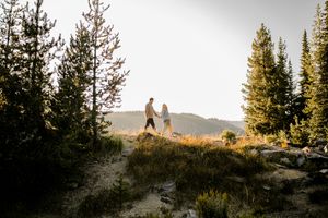 Silhouetted couple standing together on a mountain trail at sunset surrounded by evergreen trees.