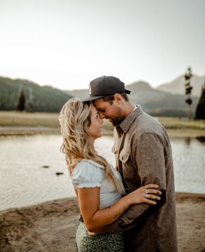 A romantic couple embraces near a mountain lake at sunset wearing casual attire.
