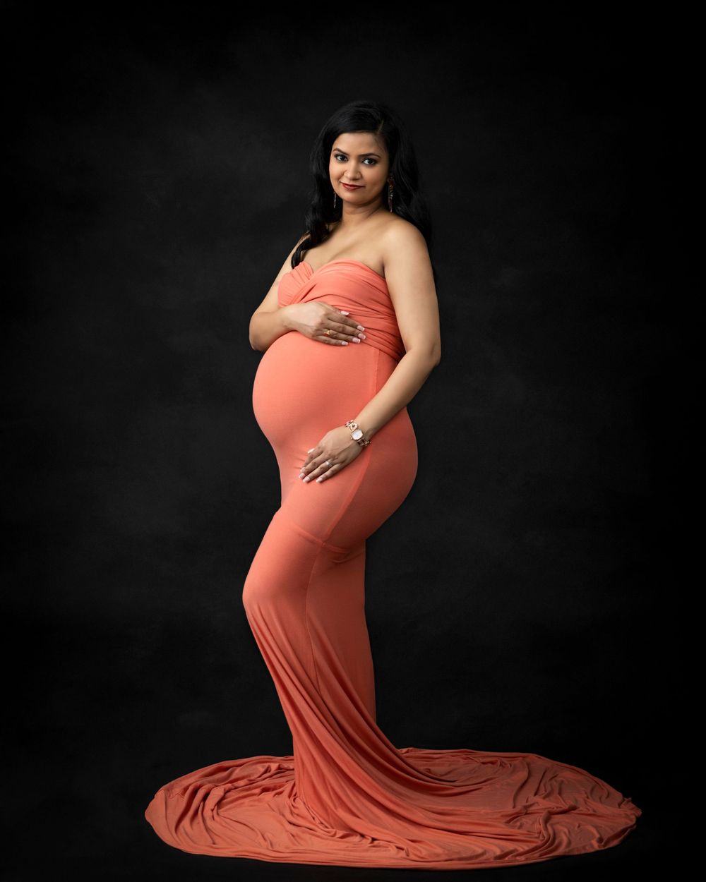 Side profile maternity portrait in a flowing coral dress on black backdrop.