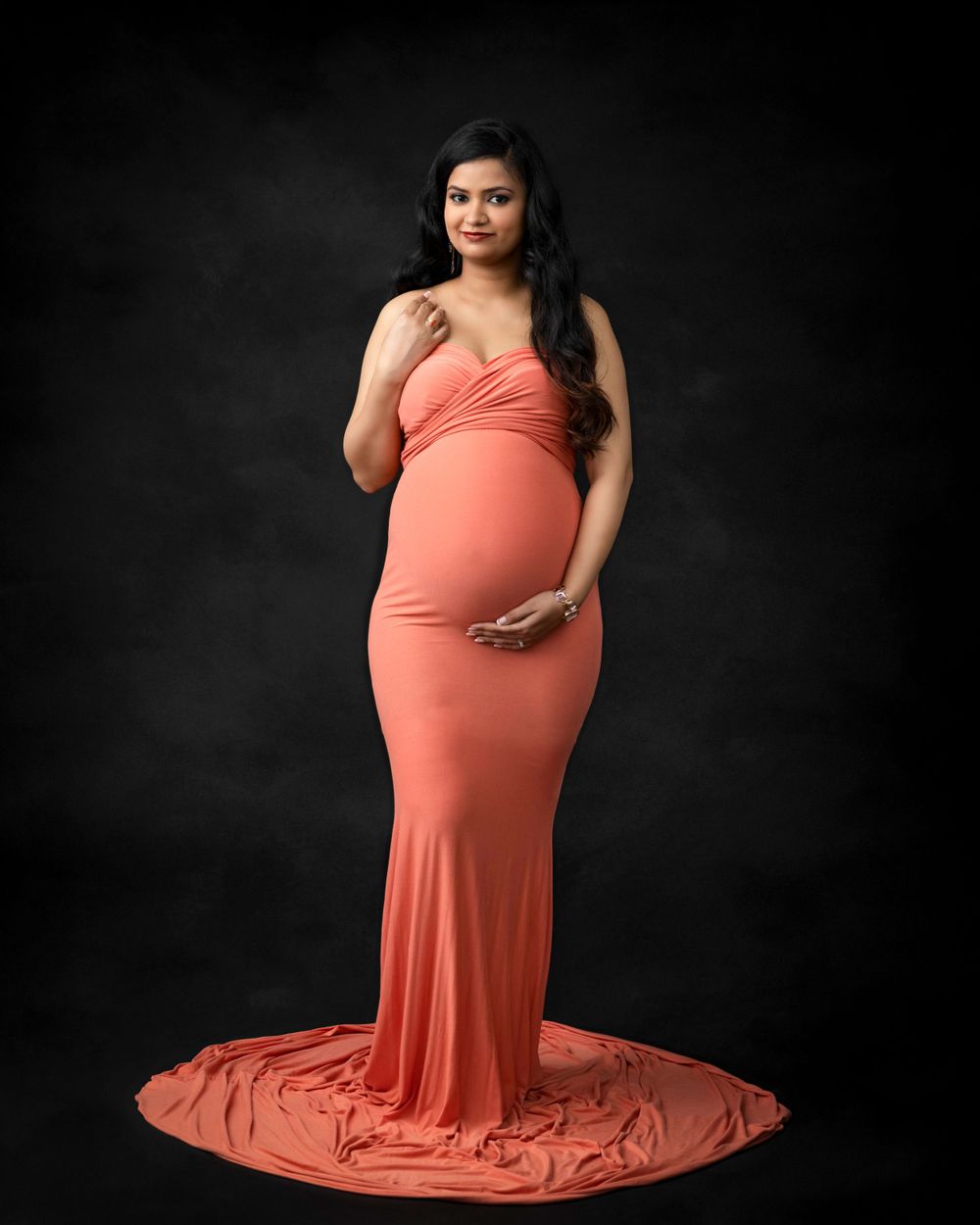 A maternity photoshoot featuring a coral floor-length gown against a dark background.