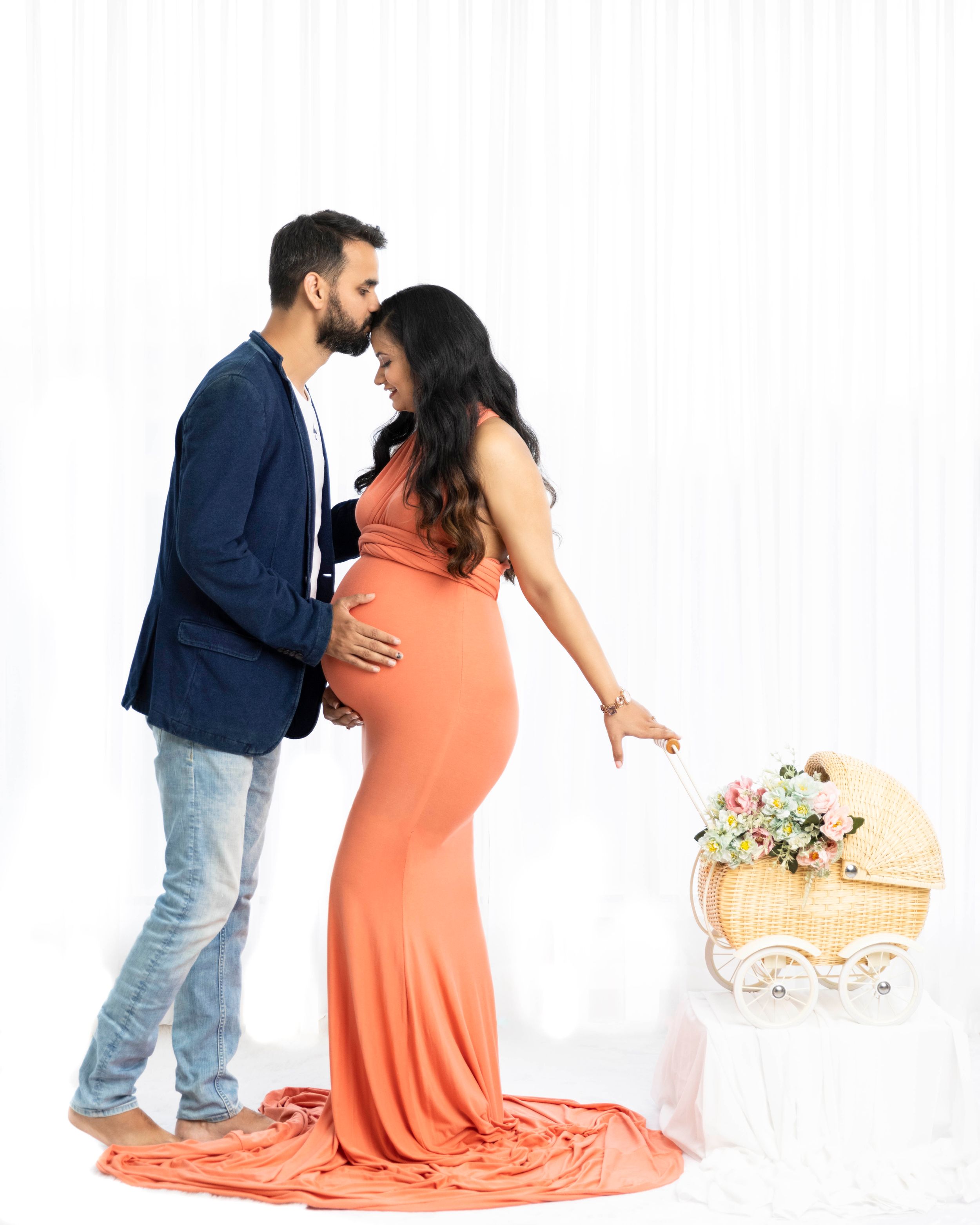 Romantic maternity photo with couple in coral dress and navy blazer next to decorative bassinet.