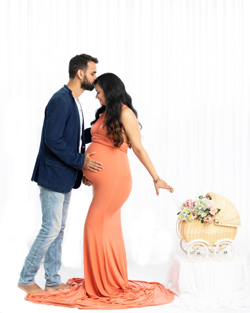 Romantic maternity photo with couple in coral dress and navy blazer next to decorative bassinet.