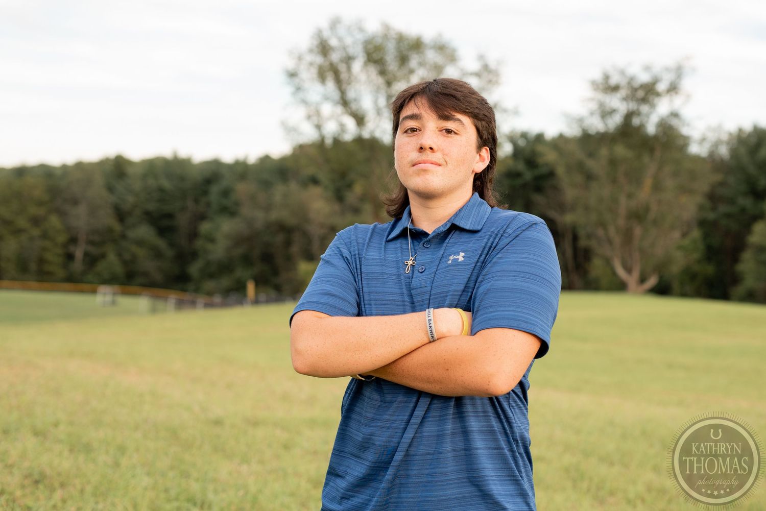A person in a blue polo shirt poses with arms crossed in a grassy field with trees in the background.