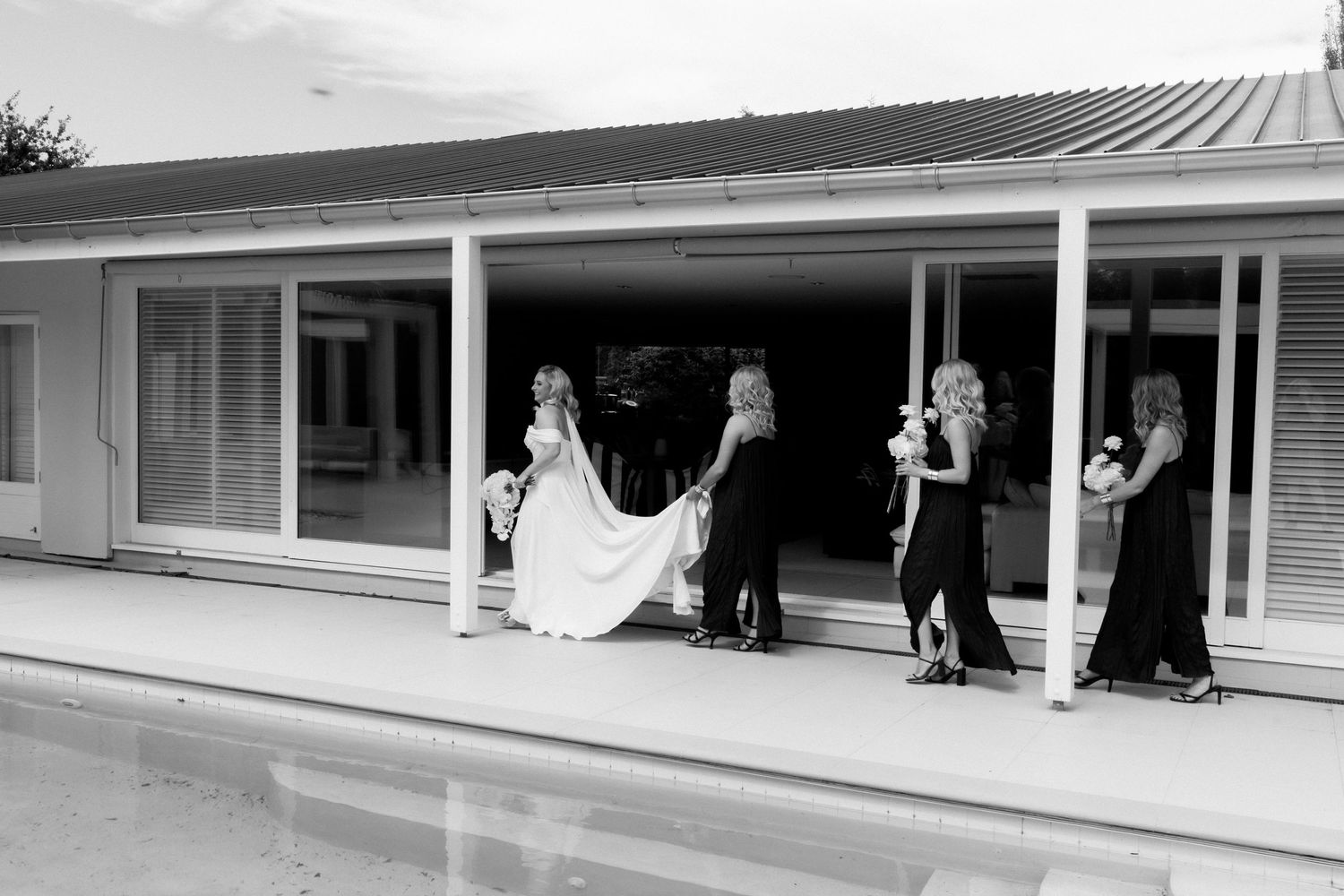 Wedding party helps with dress train outside modern building in black and white photo.