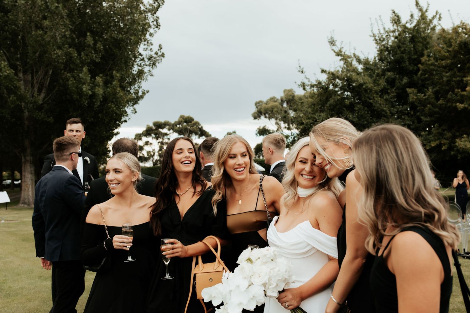 Wedding party in black dresses laughing together on grassy lawn.