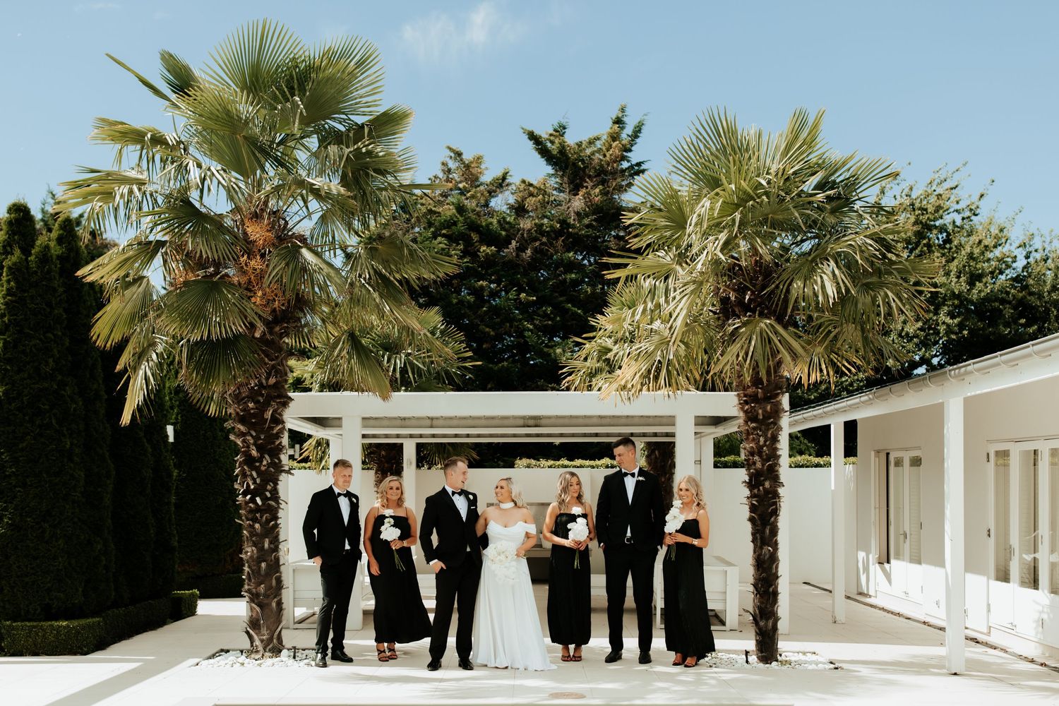 Wedding party in black and white attire poses under pergola by pool.