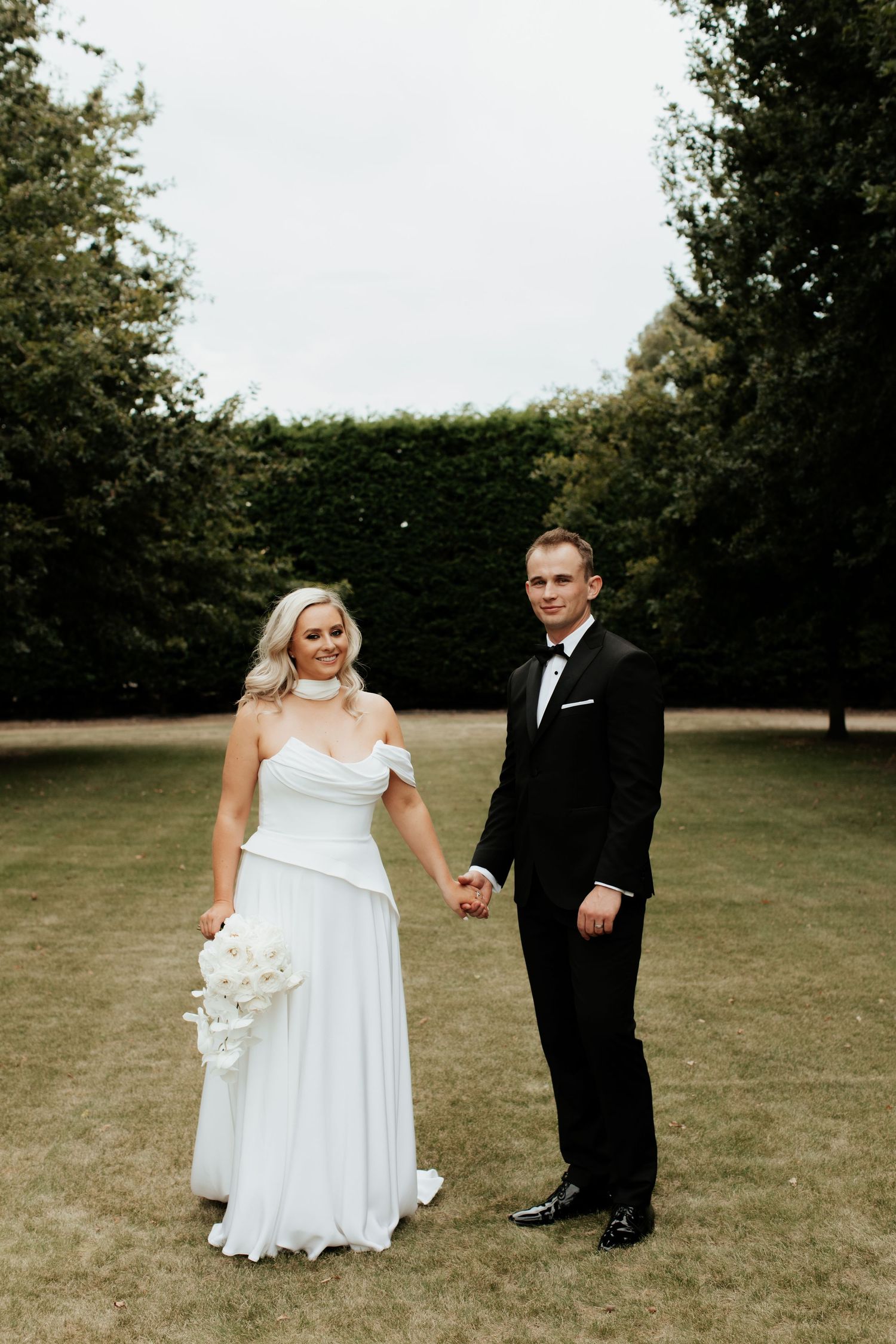 A bride in a flowing white gown stands with her groom in a black tuxedo on a grassy lawn surrounded by trees.