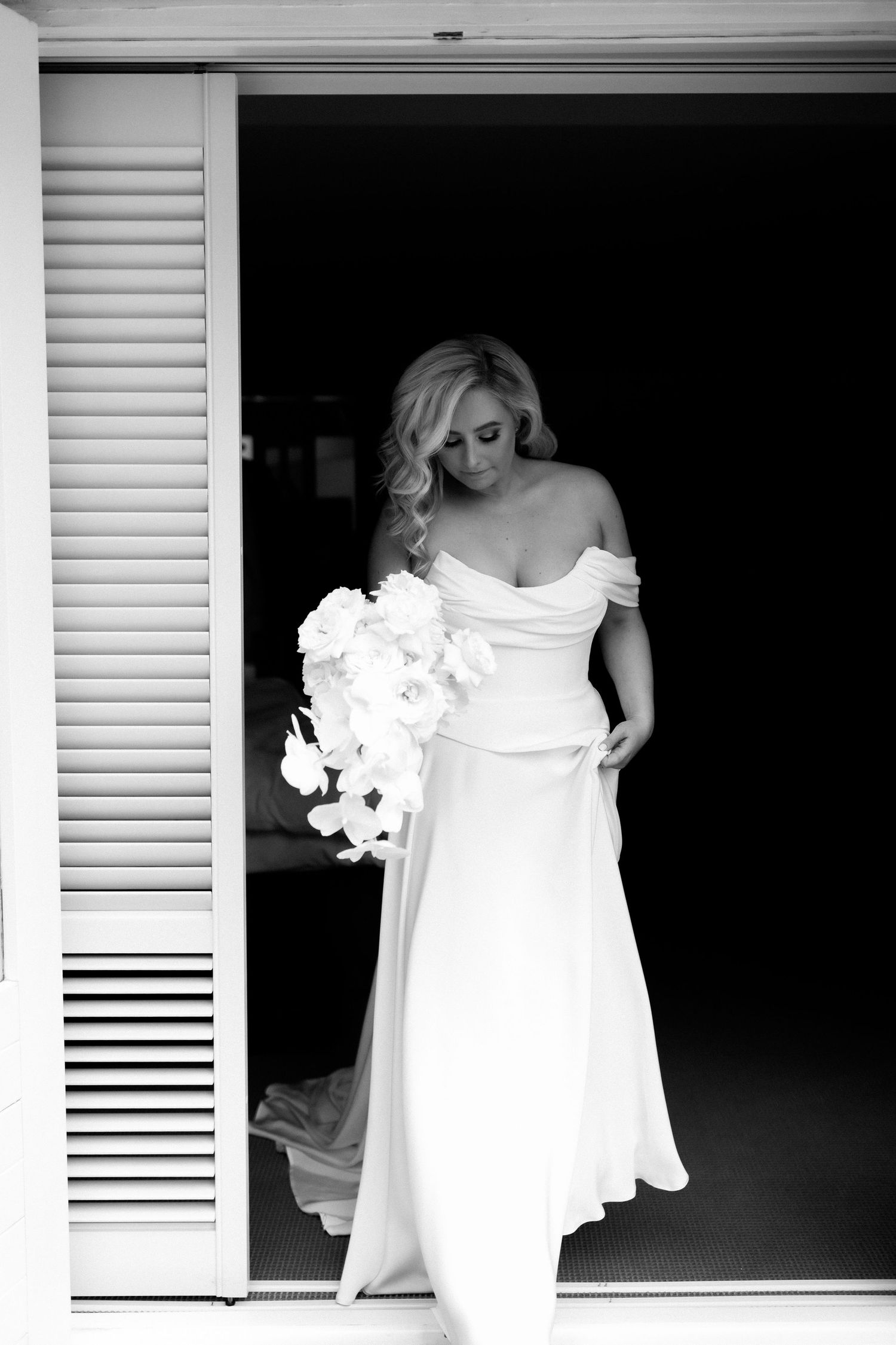 Black and white photo of bride in off-shoulder gown holding white bouquet.