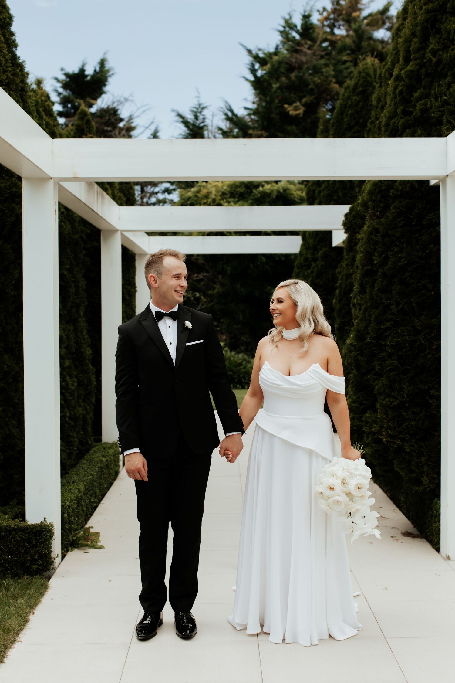 Newlyweds share romantic moment under white pergola surrounded by greenery.