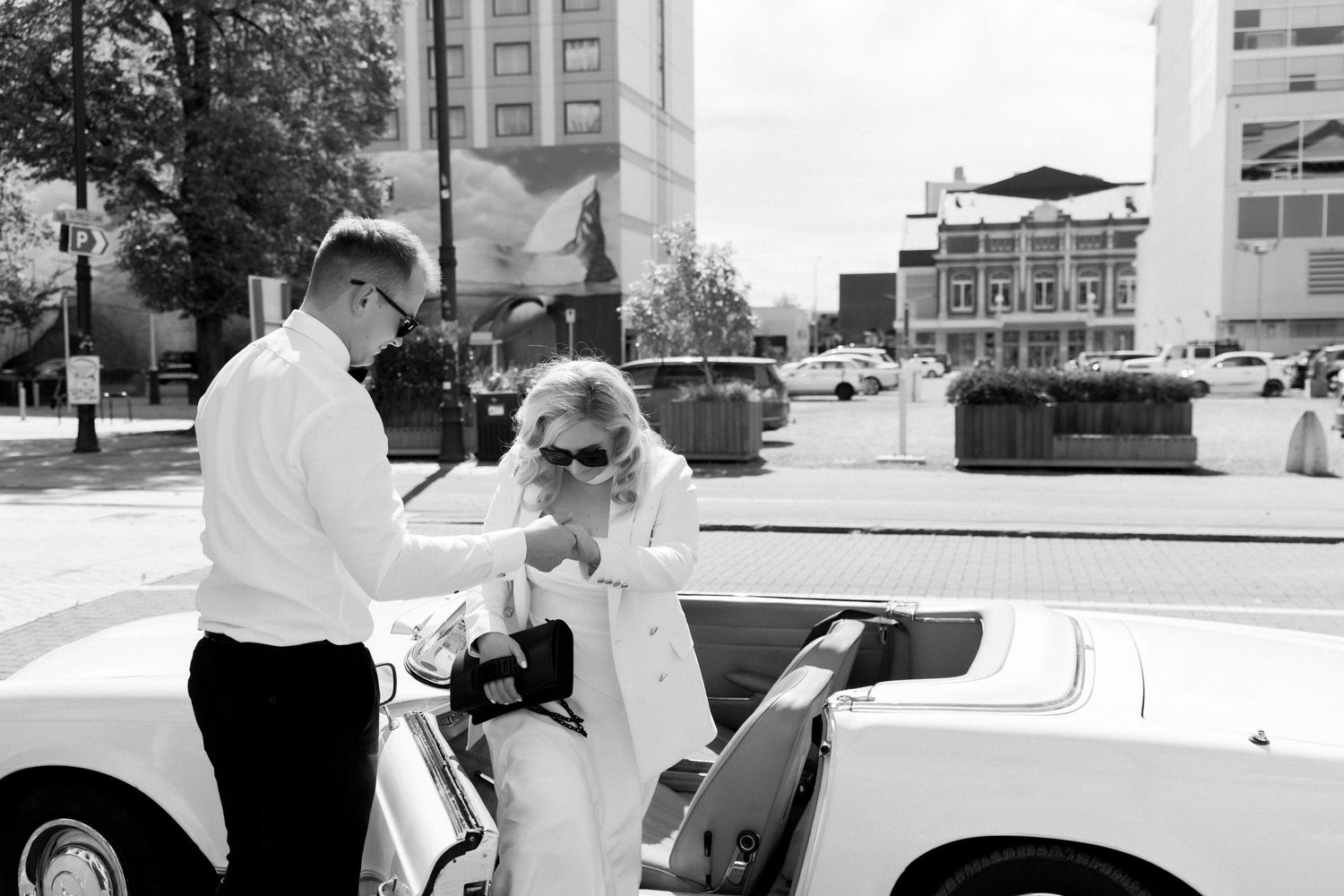 Black and white photo of wedding couple with vintage Mercedes convertible in urban setting.