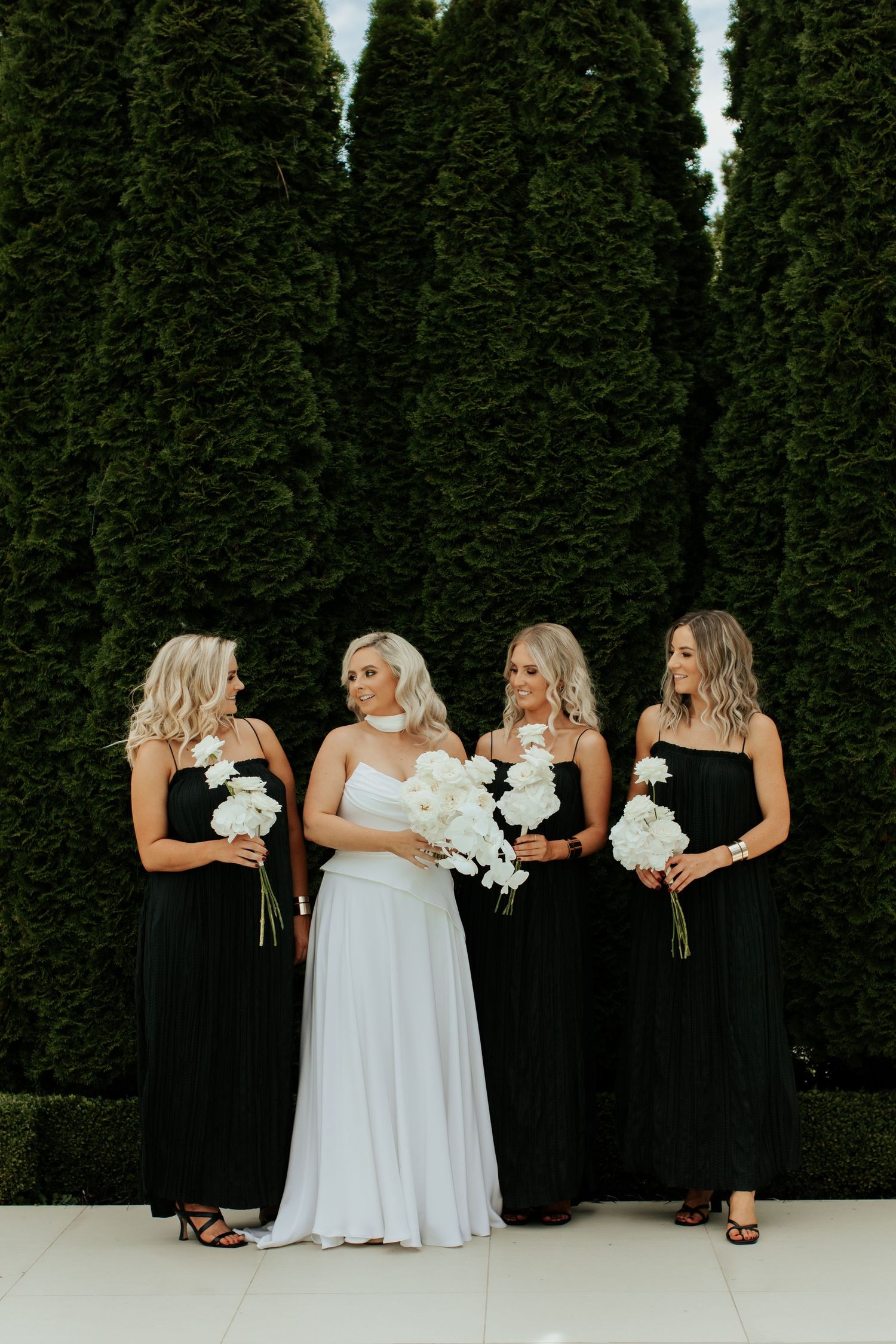 Wedding party in white and black dresses pose against tall cypress trees.