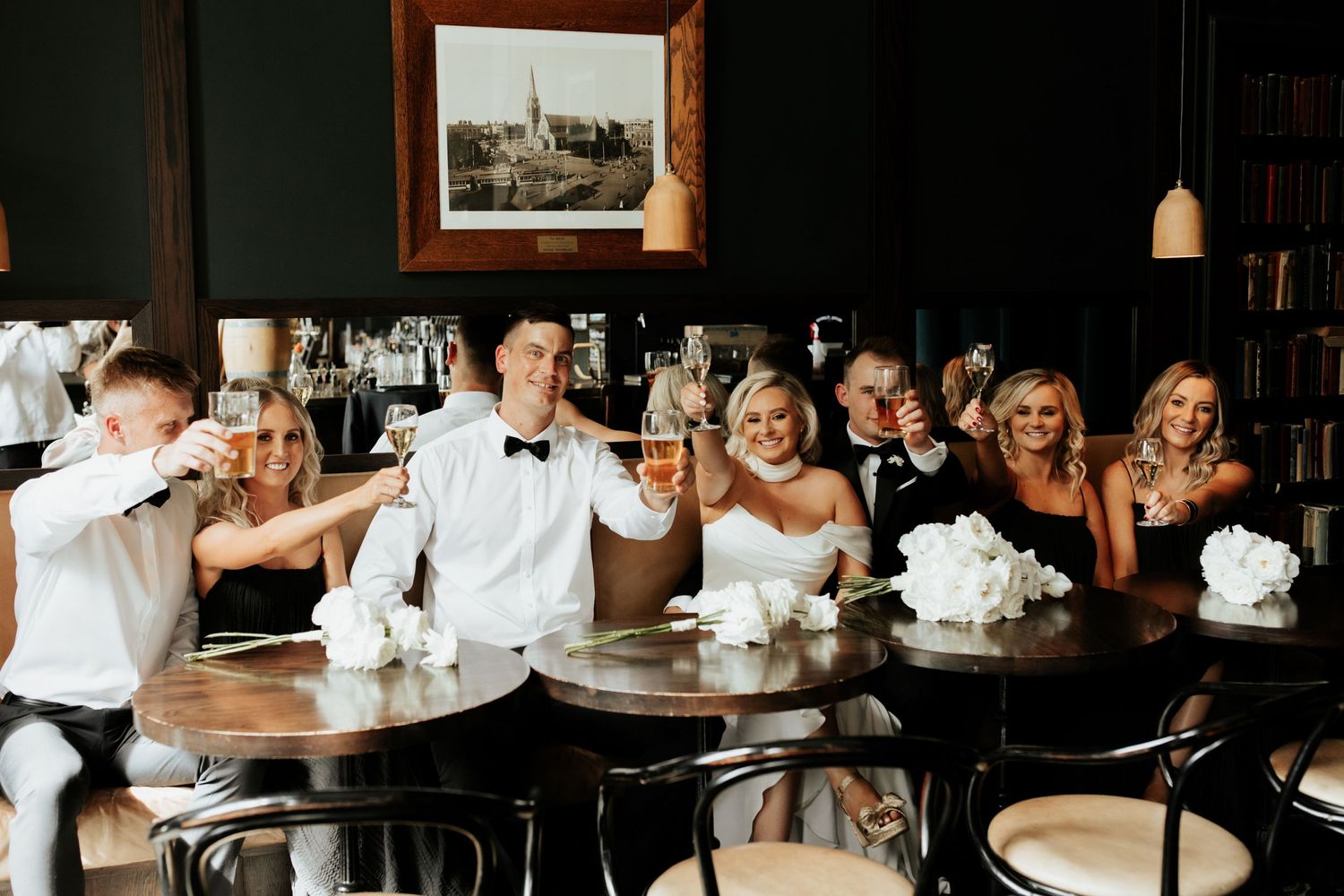 Wedding party celebrates with champagne toasts at a long wooden table decorated with white floral centerpieces.