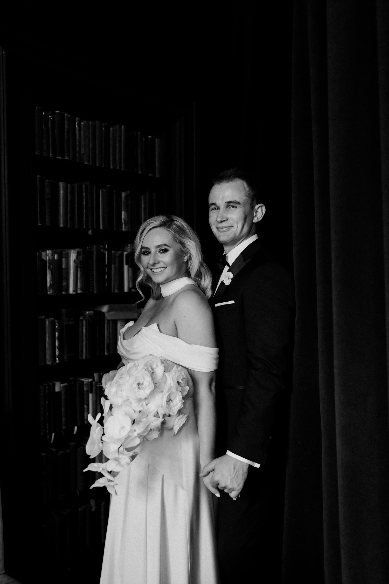 Romantic wedding portrait in front of a dark bookshelf with off-shoulder white gown and classic tuxedo.