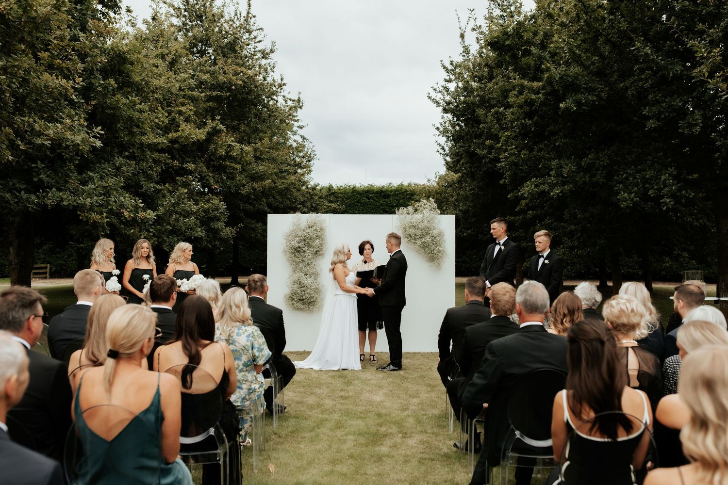 Outdoor wedding ceremony with white backdrop and guests seated on lawn.