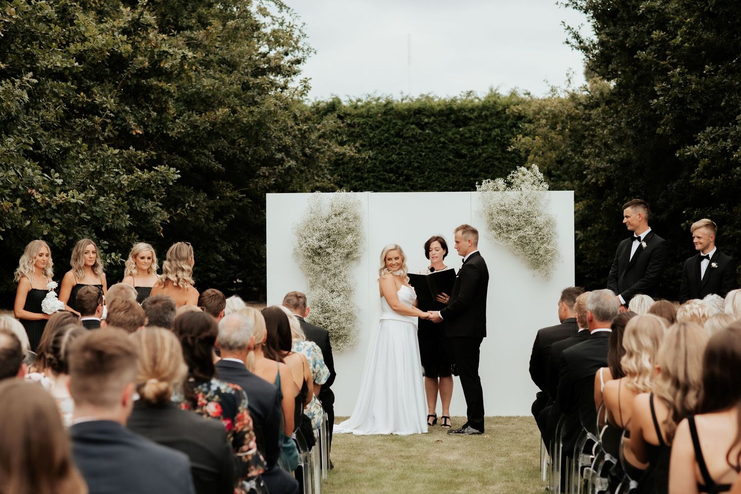 Garden wedding ceremony with white floral backdrop and guests in formal attire.