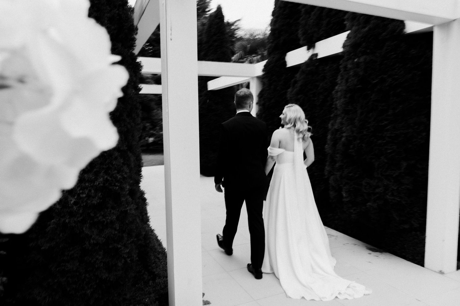 Artistic black and white photo of couple walking under white pergola structure.