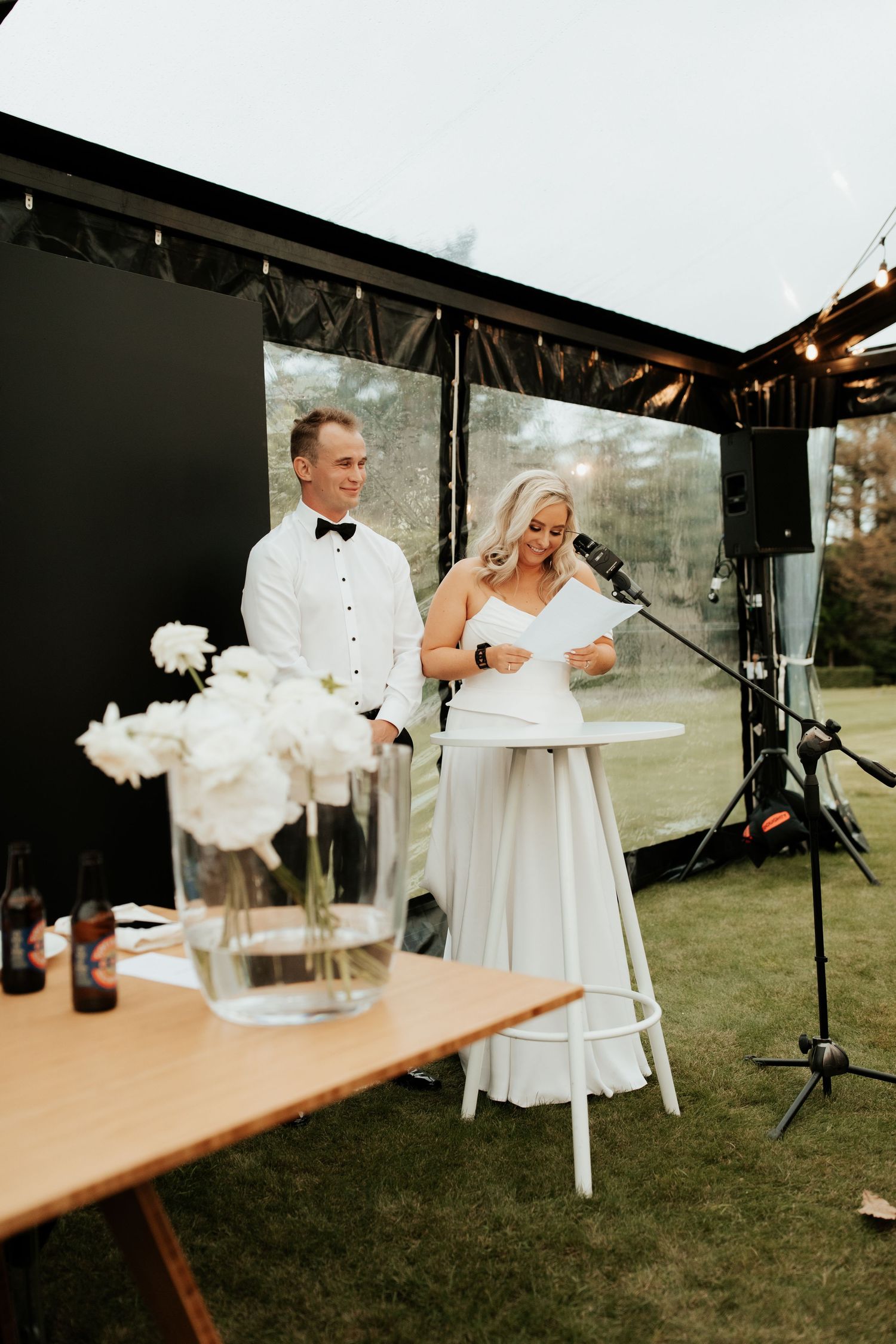 Wedding couple delivers speeches during outdoor reception with white floral centerpieces.