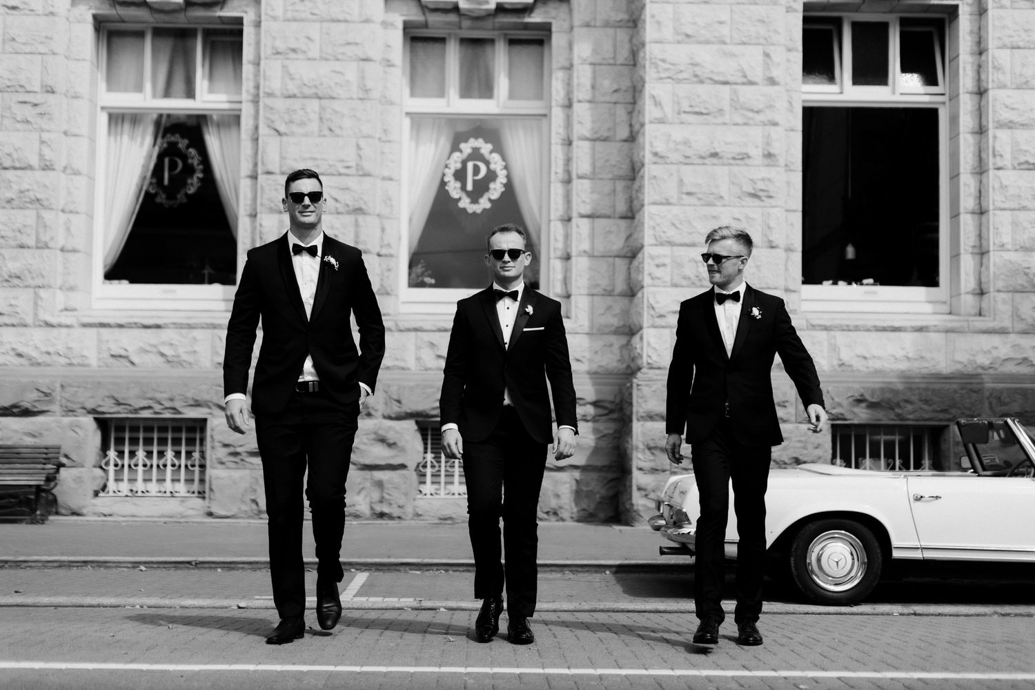 Three groomsmen in classic black tuxedos walking outside a stone building.