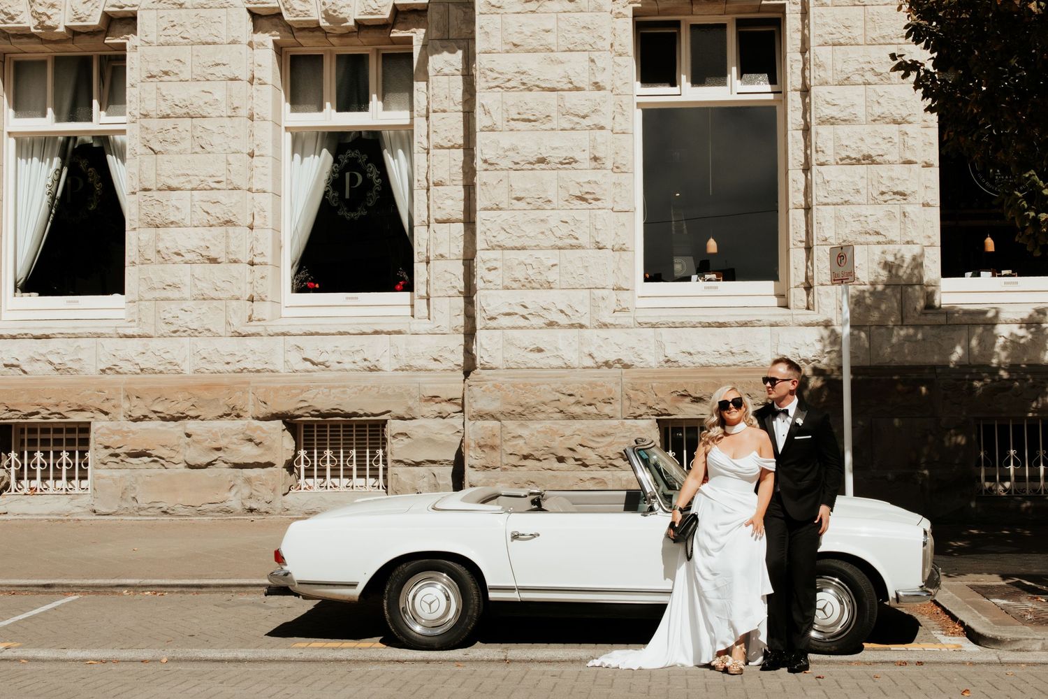 Wedding photo with classic white Mercedes-Benz convertible against stone building backdrop.