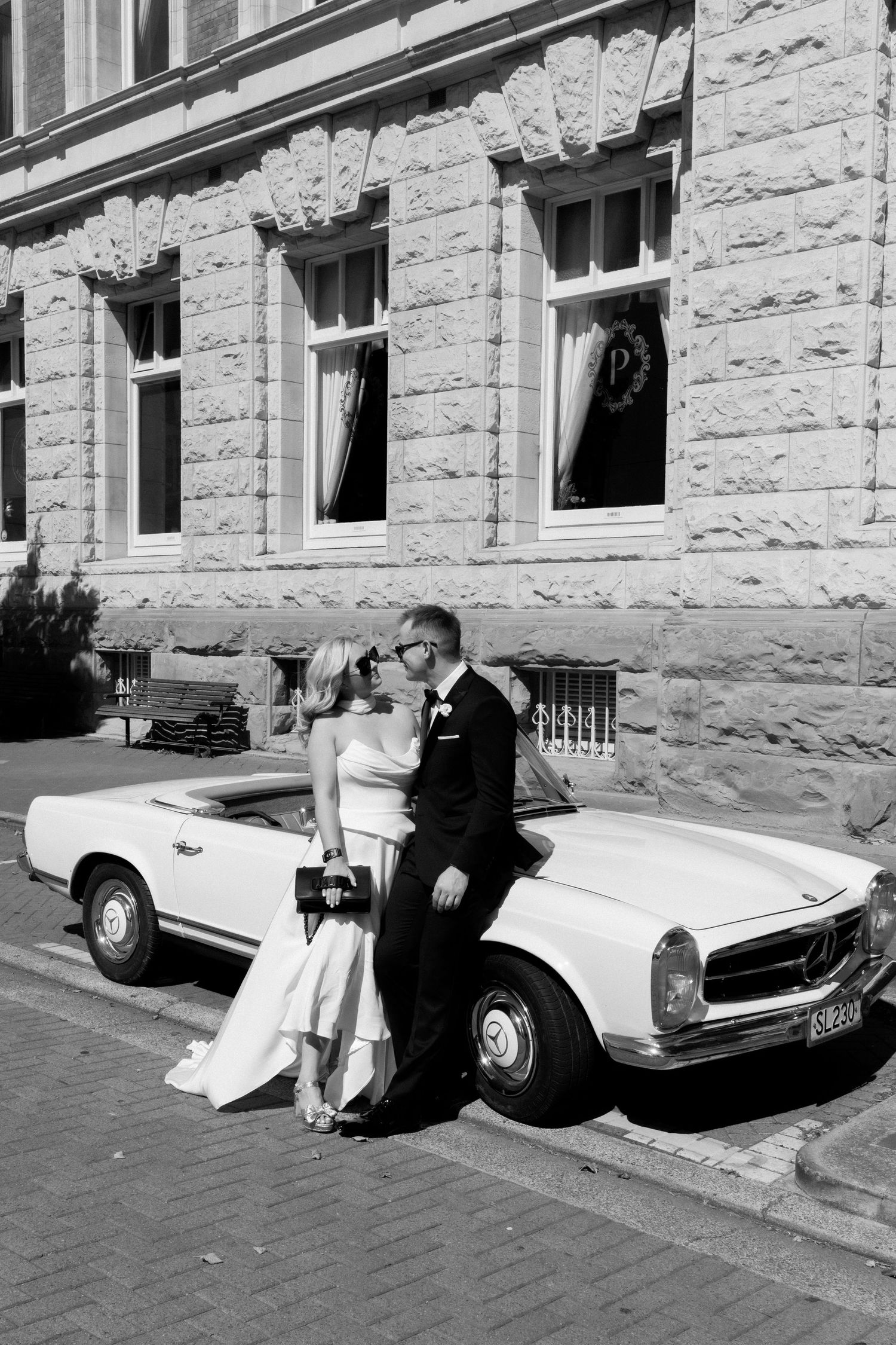 A vintage classic Mercedes convertible parked outside a stone building with a couple in wedding attire posing beside it.