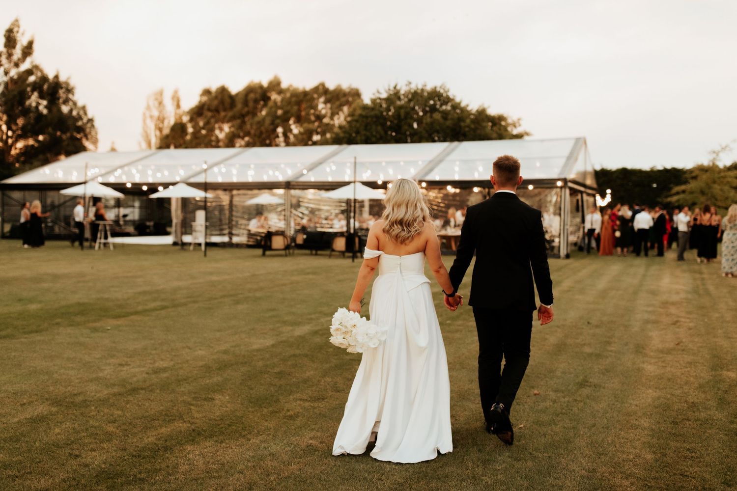 Couple walking towards outdoor wedding reception tent on a grassy field.