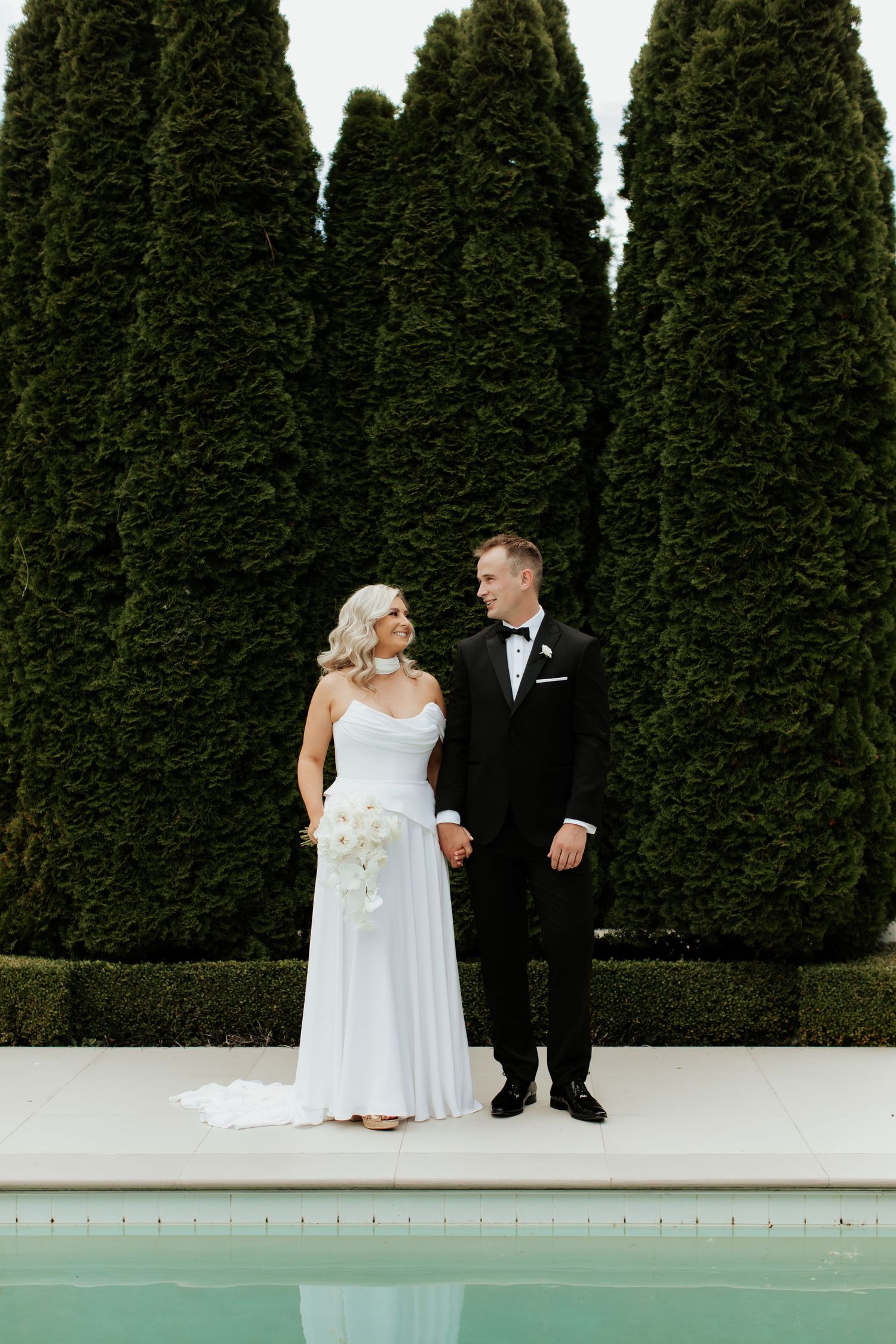 Happy couple poses against tall cypress trees near a pool in formal wedding attire.