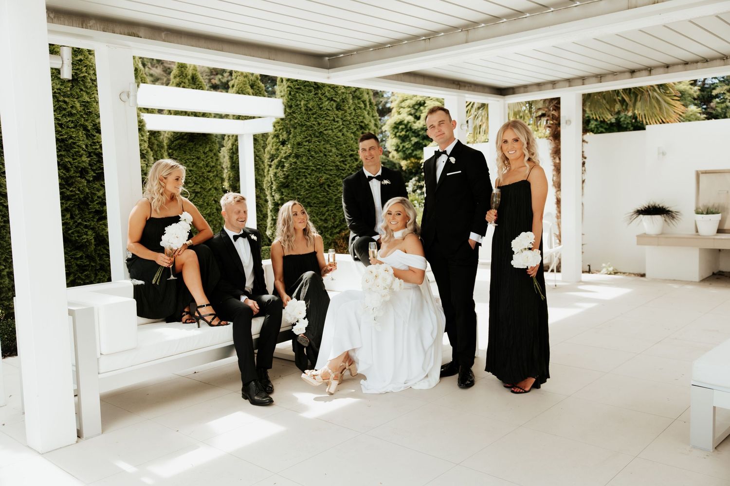 Wedding party gathered under white pergola for casual group photos.