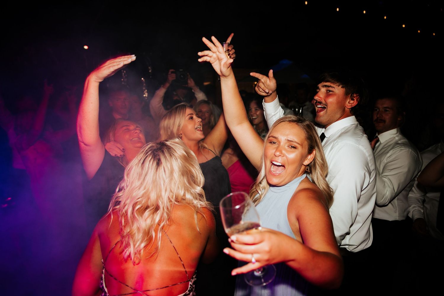 Wedding guests dancing and celebrating on crowded dance floor with purple mood lighting.