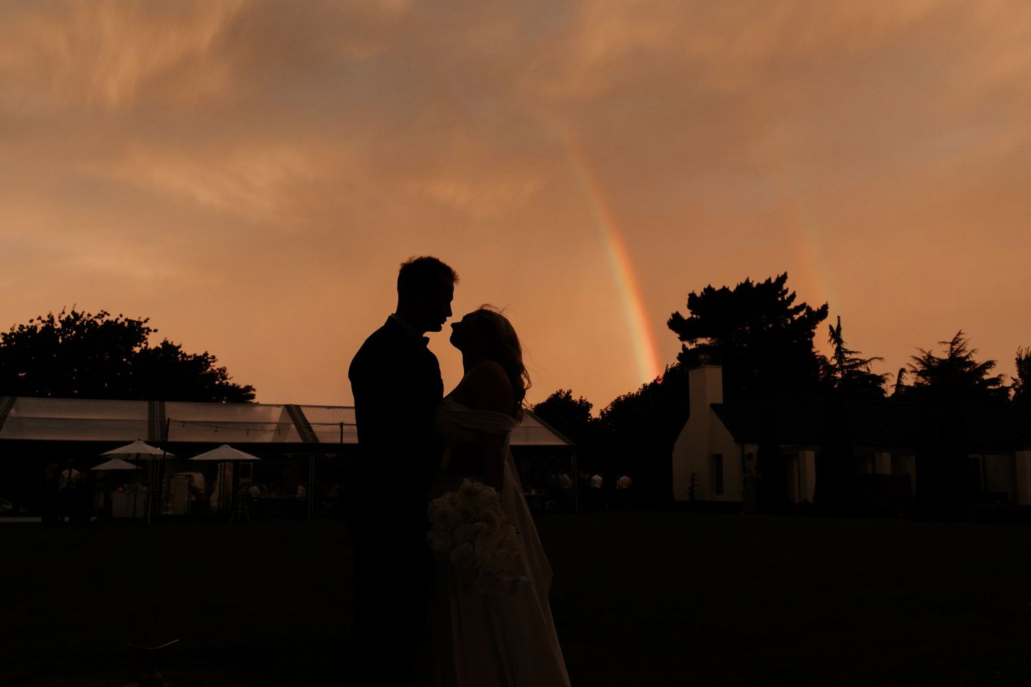 Silhouette of a couple embracing at sunset with a rainbow in the background during their outdoor wedding.