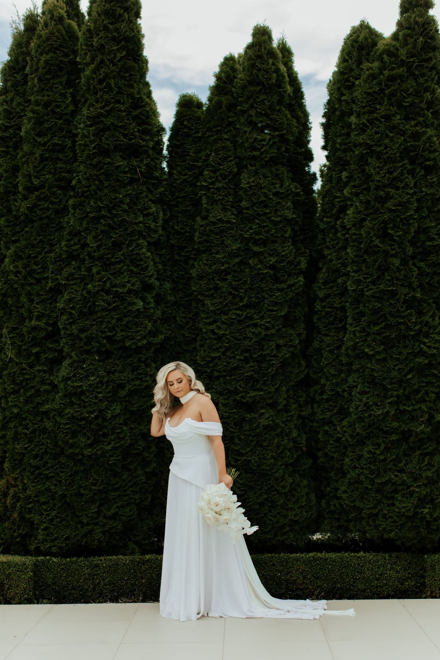Elegant bride in flowing white off-shoulder gown poses against tall emerald cypress trees.