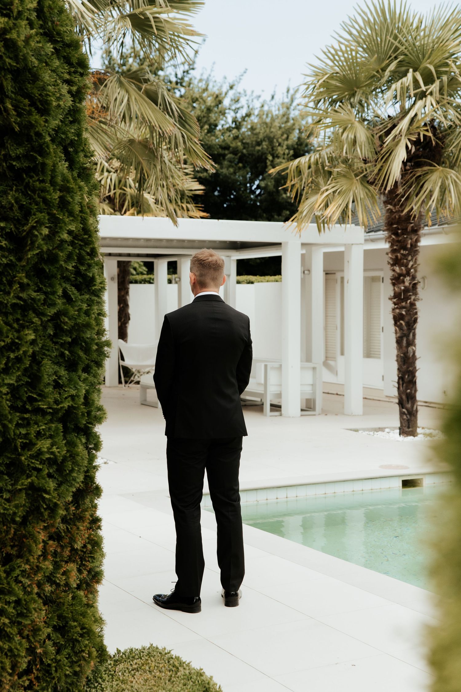 Person in formal attire standing by pool with palm trees and white pergola.