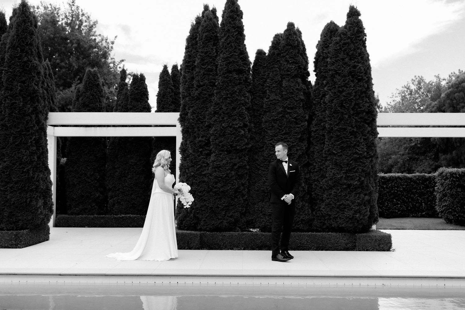 Black and white wedding photo of couple standing apart by swimming pool with cypress trees.
