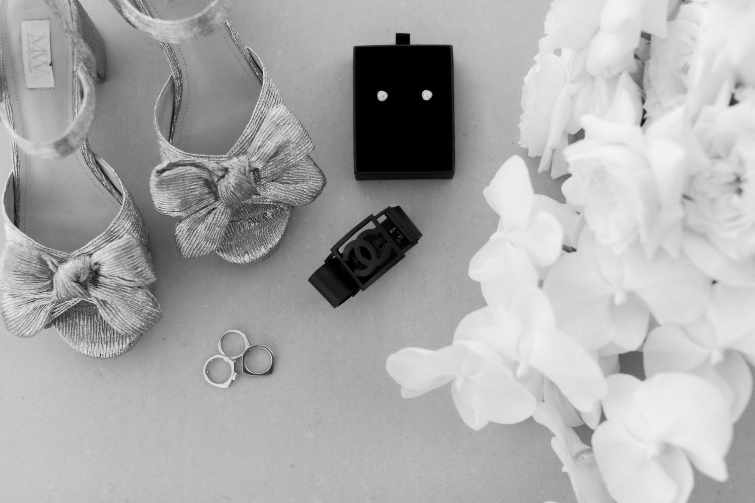Wedding accessories including silver shoes, rings, and white flowers arranged on light background.