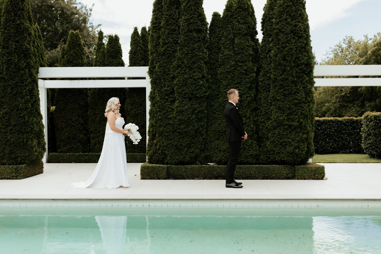 Couple in wedding attire standing separately by pool with manicured hedges.