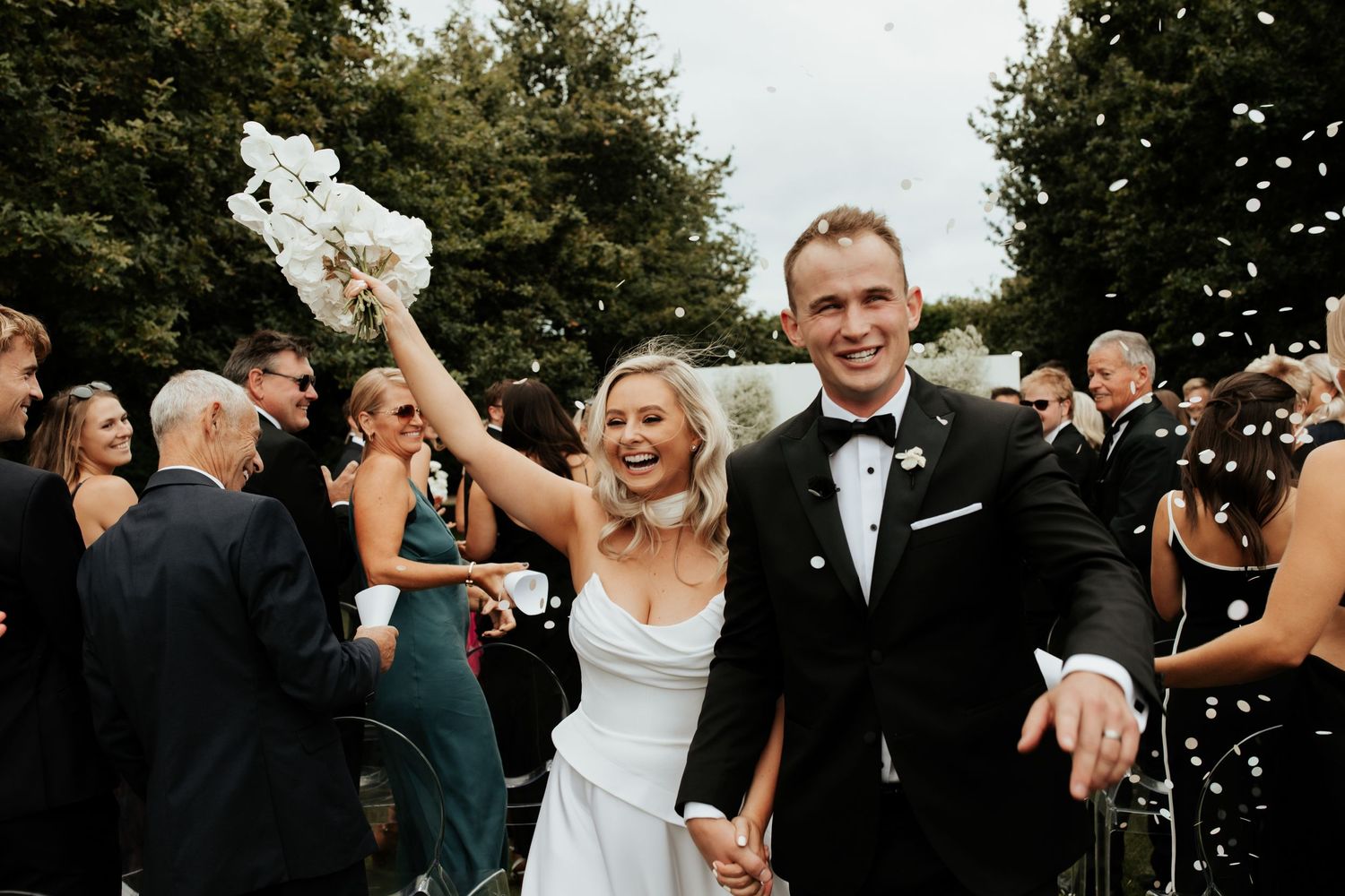 Newlyweds celebrating with guests throwing confetti in an outdoor garden setting.