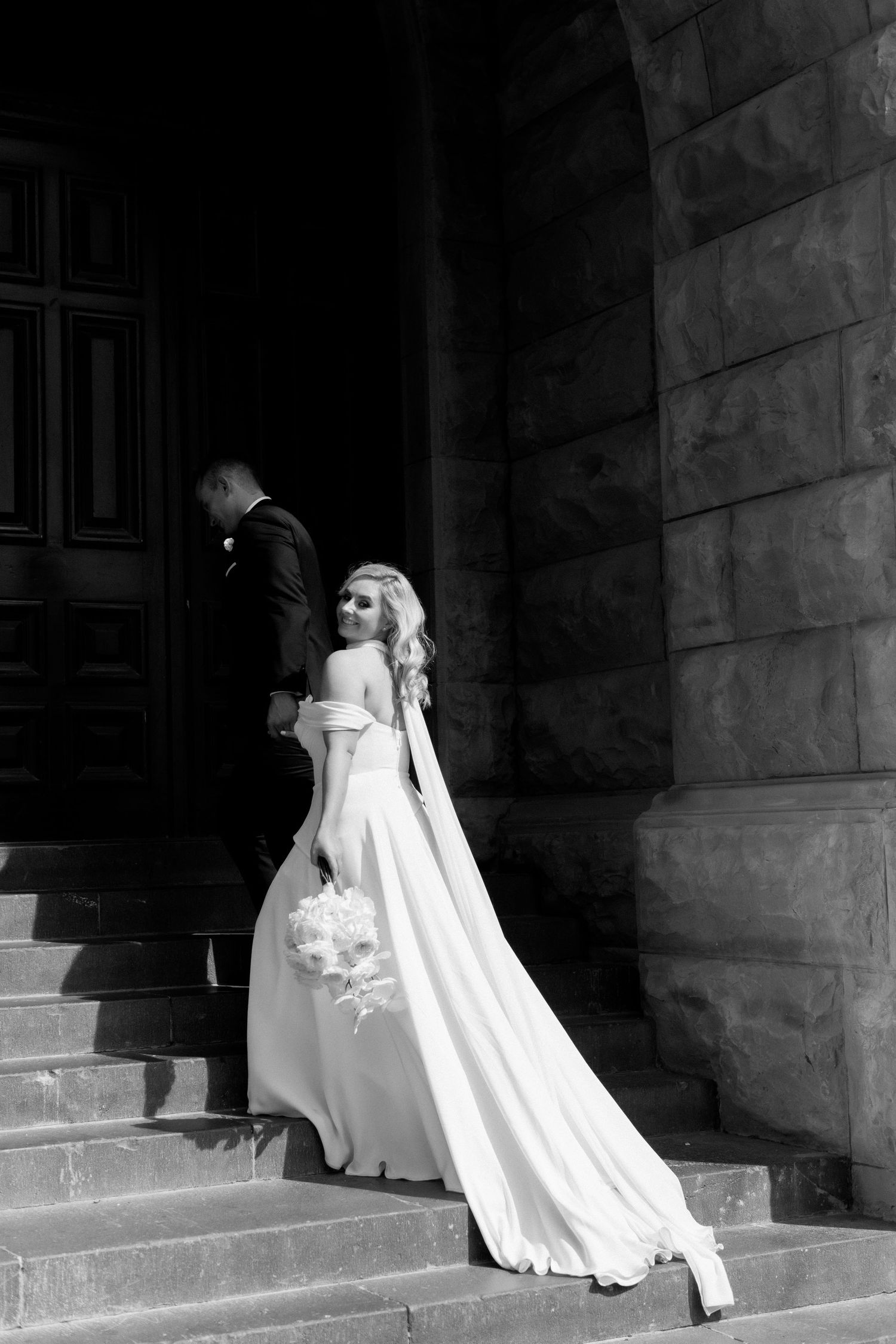 Dramatic black and white shot of a wedding couple ascending stone steps in formal attire.