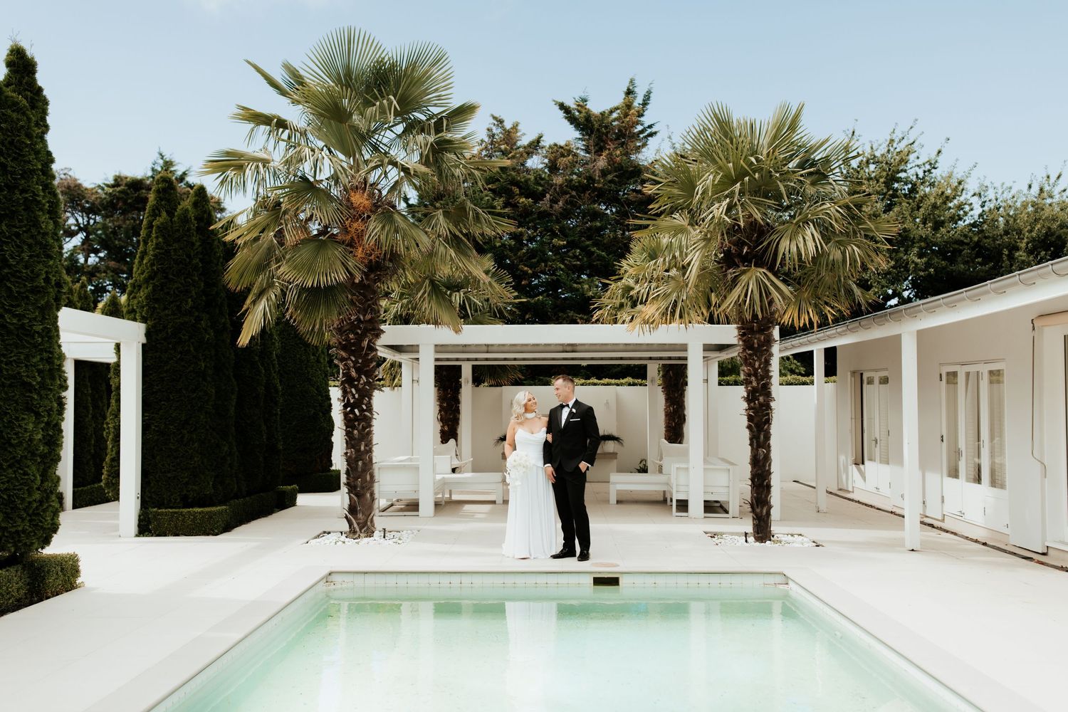 Wedding couple stands under white pergola by pool with palm trees and modern architecture.