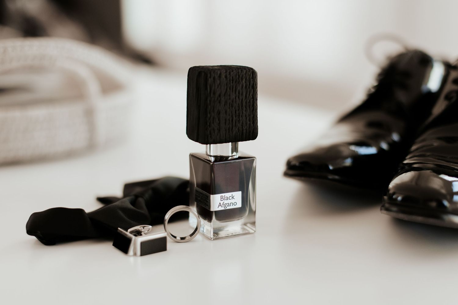 Elegant black cologne bottle with bow tie and dress shoes arranged on white surface.