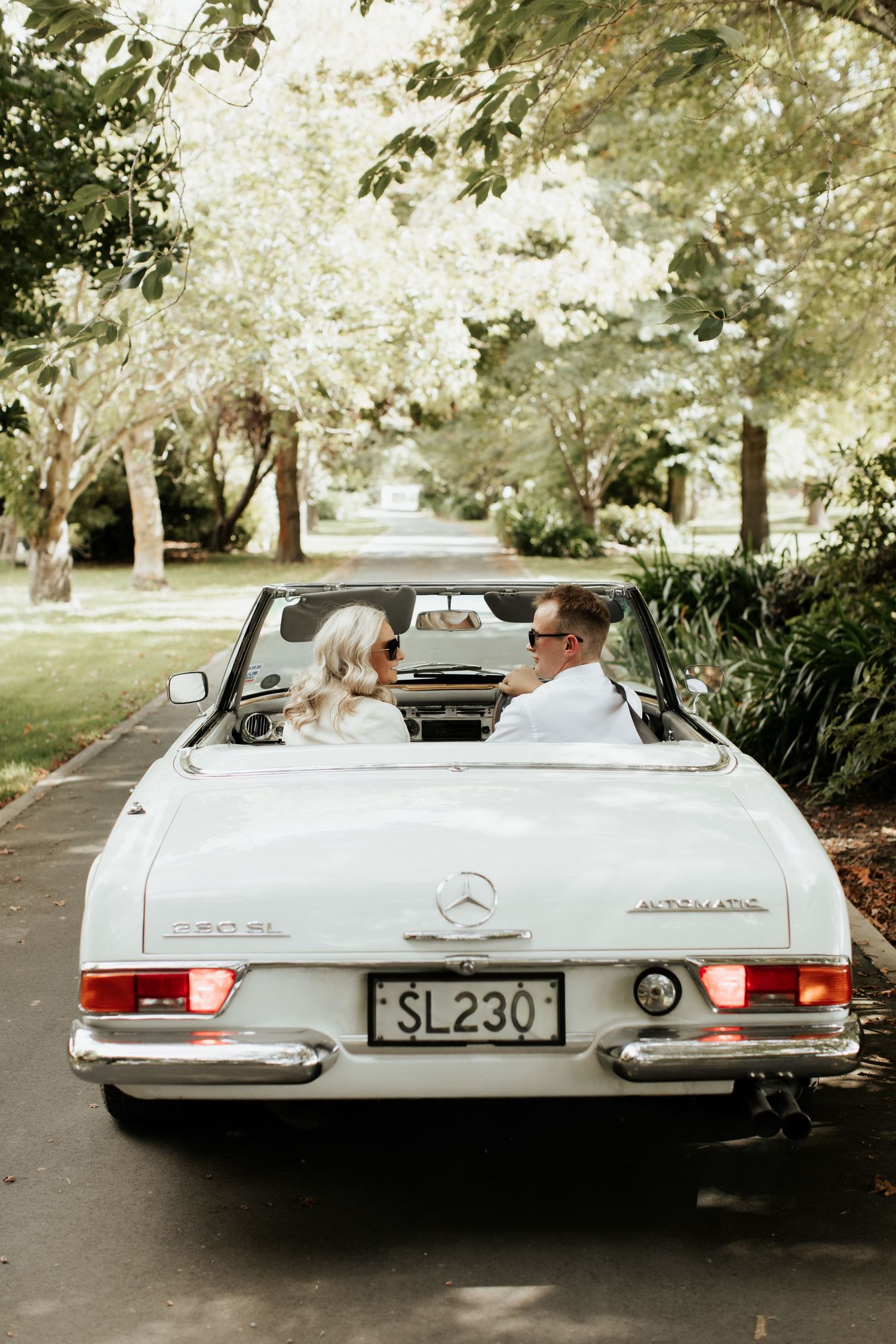 Couple driving in a classic white Mercedes-Benz convertible down a tree-lined street.