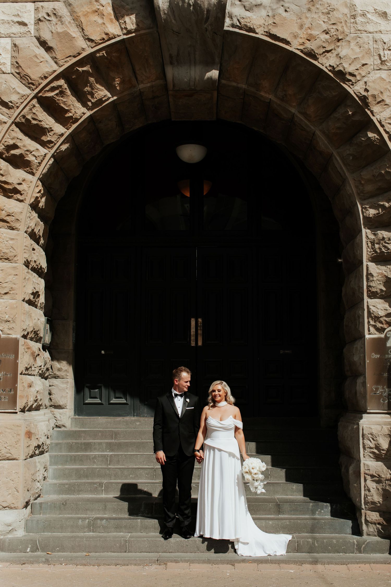 Wedding couple stands beneath a grand stone archway entrance in classic formal attire.