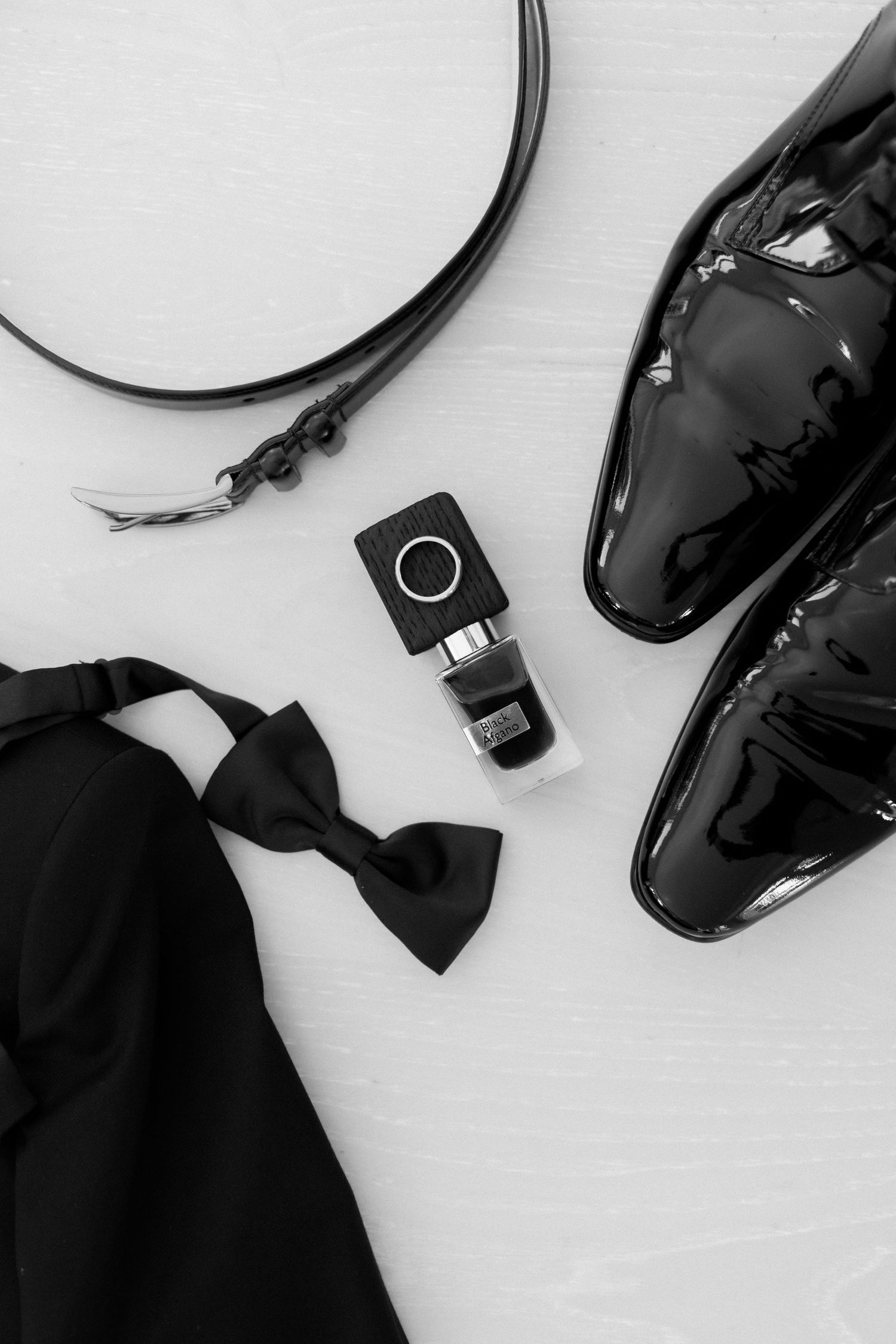 Wedding accessories including black dress shoes, bowtie and ring box on white surface.