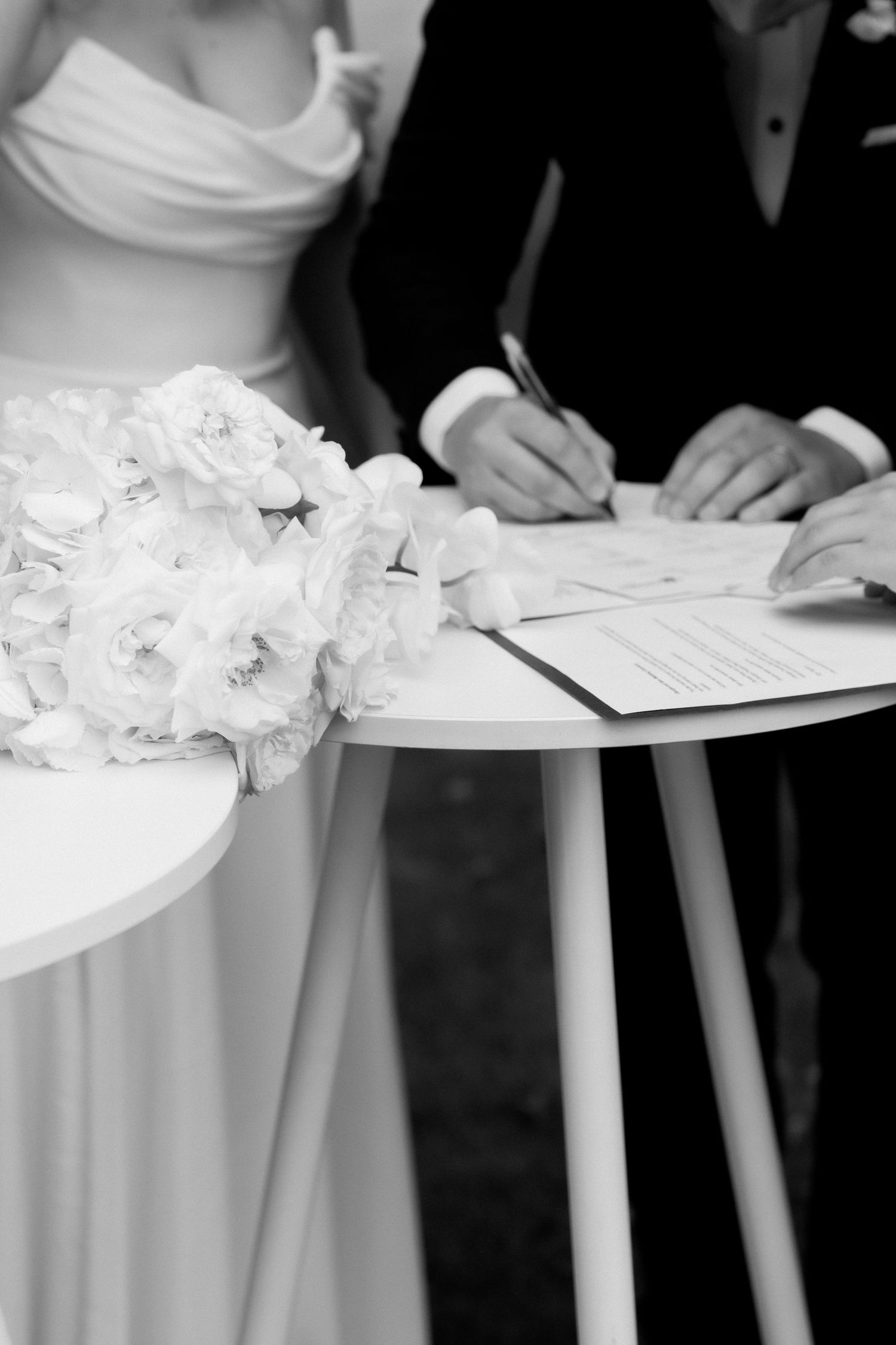 Close-up black and white shot of signing wedding documents with white bouquet on table.
