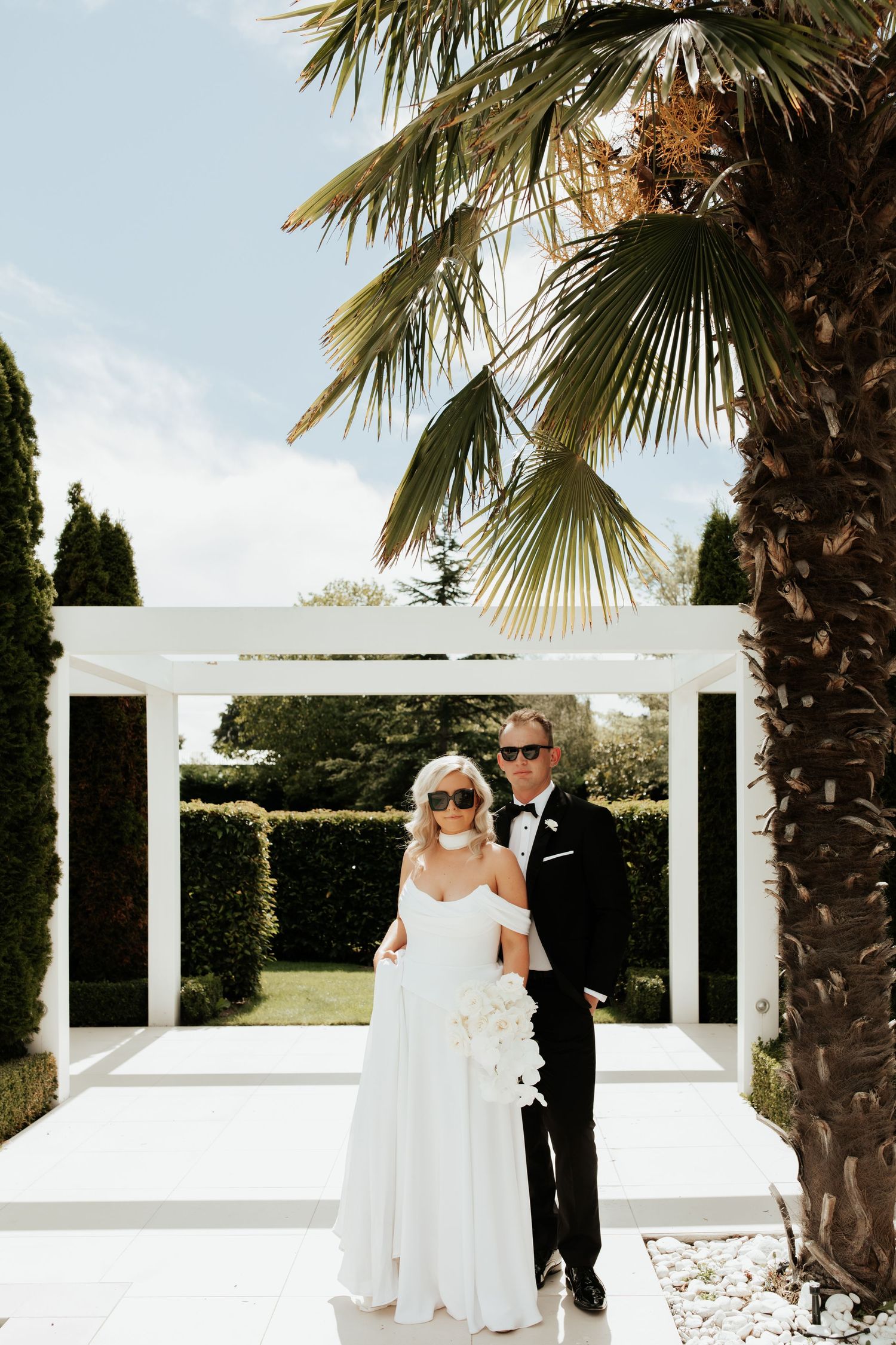 Couple poses under white pergola with palm trees in tropical garden setting.