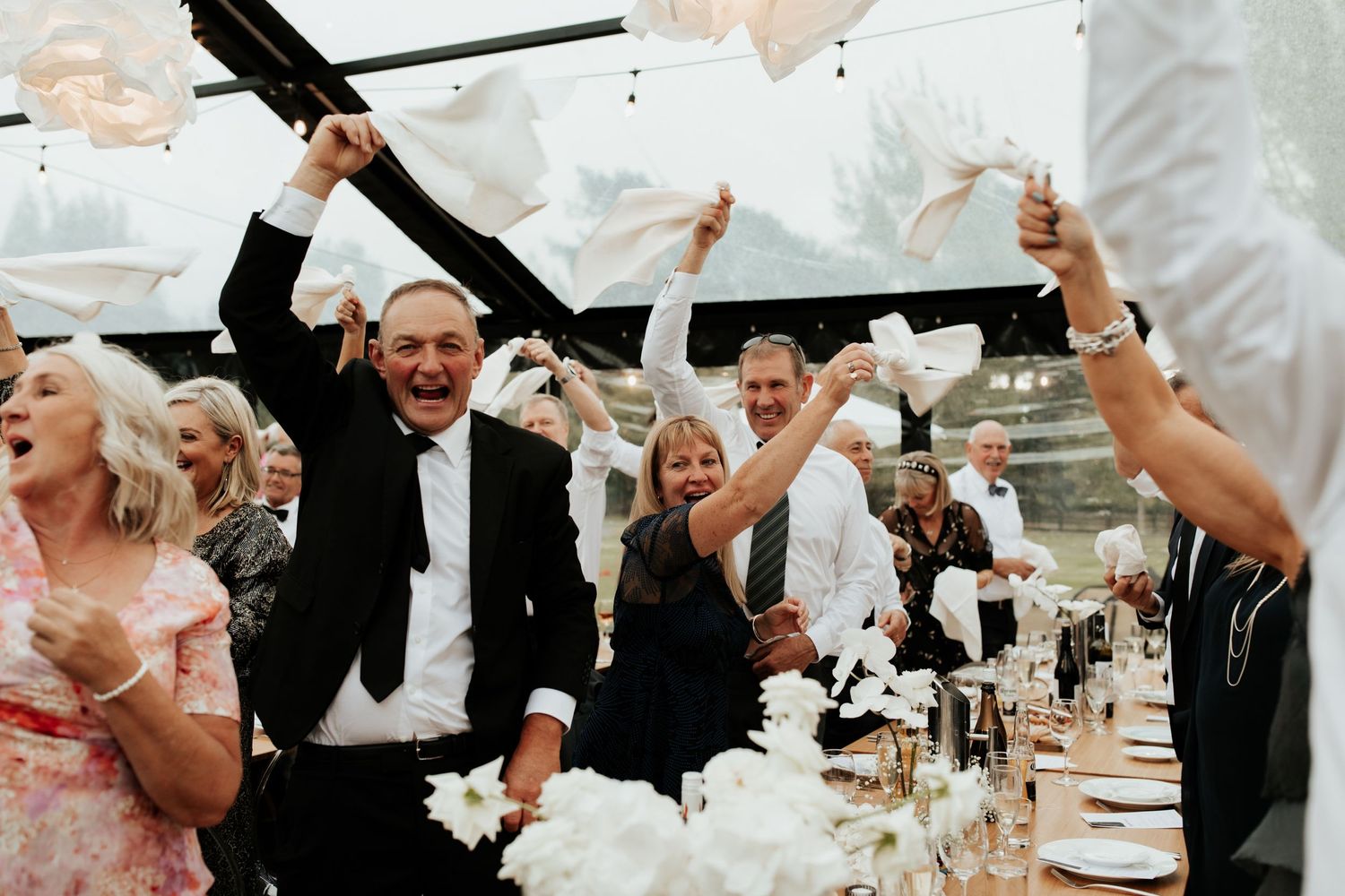 Wedding reception guests celebrate with raised hands under a transparent tent at sunset.