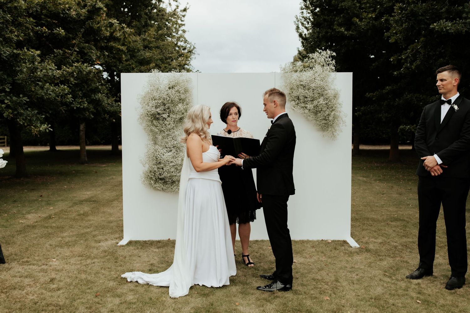 Wedding ceremony taking place outdoors with white floral backdrop and officiant performing the service.